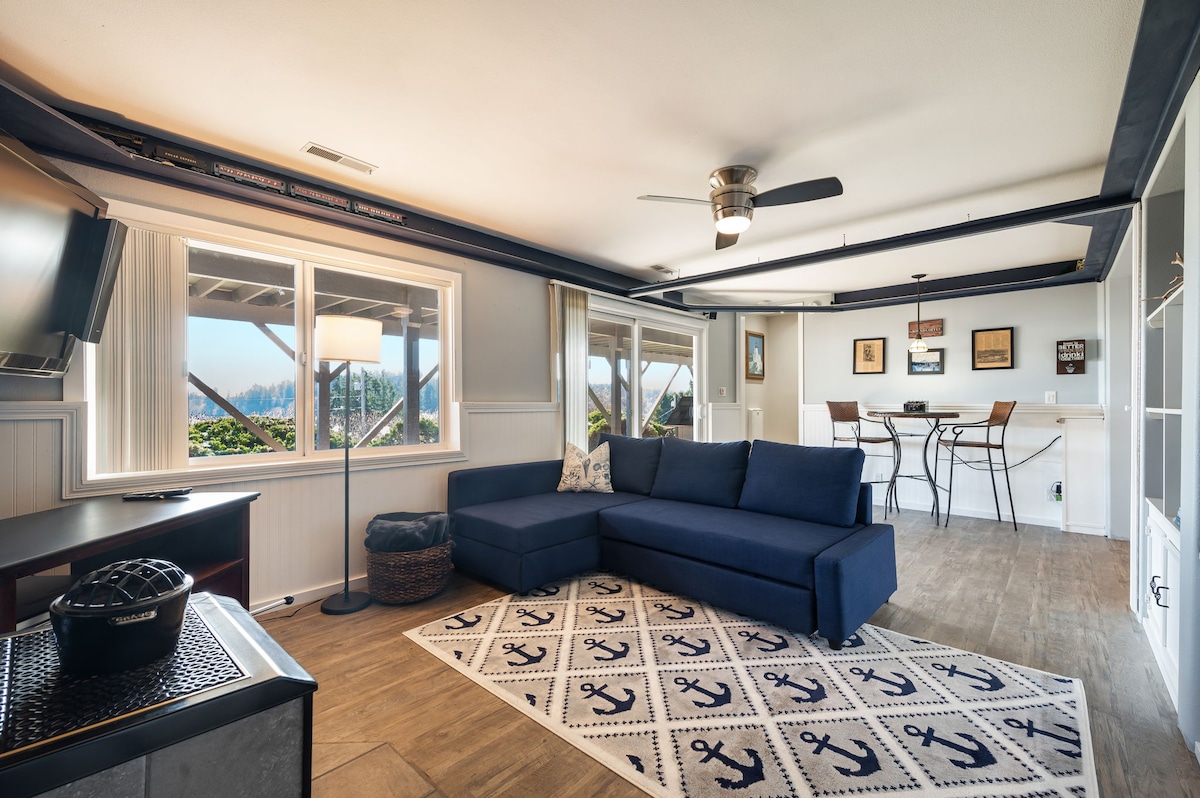 A bright living area features large windows displaying views of the surroundings. A navy blue sectional sofa faces a flat-screen TV, while a small dining table for four is positioned near the window. Light streams in, creating a spacious and airy atmosphere.