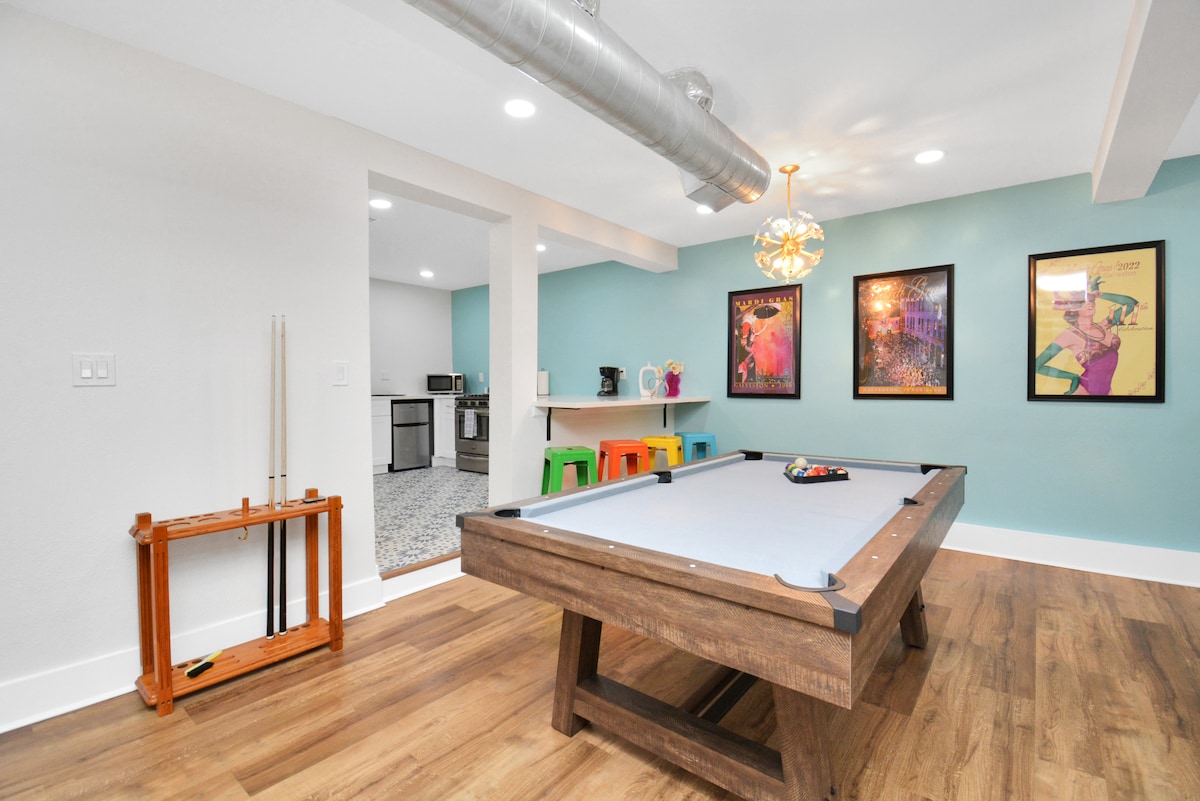 An entertainment room features a wooden pool table at the center, with colorful framed artwork adorning the walls. A vibrant light fixture hangs from the ceiling, and a kitchenette is visible in the background. Seating is provided by bright stools in varying colors.