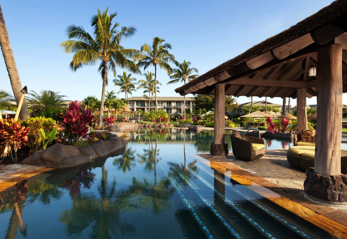 The outdoor pool area is surrounded by lush tropical plants and palm trees, with clear blue water reflecting the sky. Comfortable seating spaces are located under a shaded structure, providing a relaxing view of the resort and vibrant landscaping.