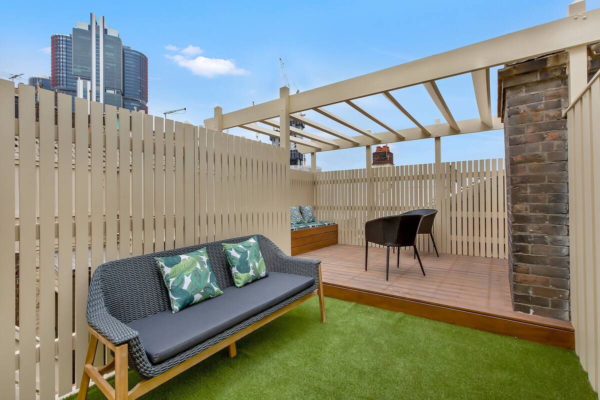 A private rooftop terrace is presented with a combination of seating options, including a stylish sofa adorned with decorative cushions and a dining area featuring dark wicker chairs. The terrace is enclosed by a light-colored fence, offering views of the surrounding urban landscape.