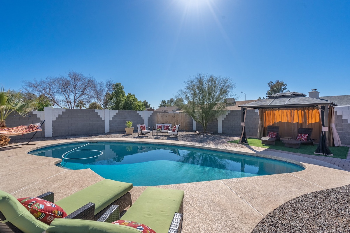 A spacious outdoor area features a heated saltwater pool surrounded by a sunbathing space and greenery. Lounge chairs are positioned for relaxation, while a shaded cabana offers comfortable seating. The clear blue sky enhances the inviting atmosphere of the backyard.