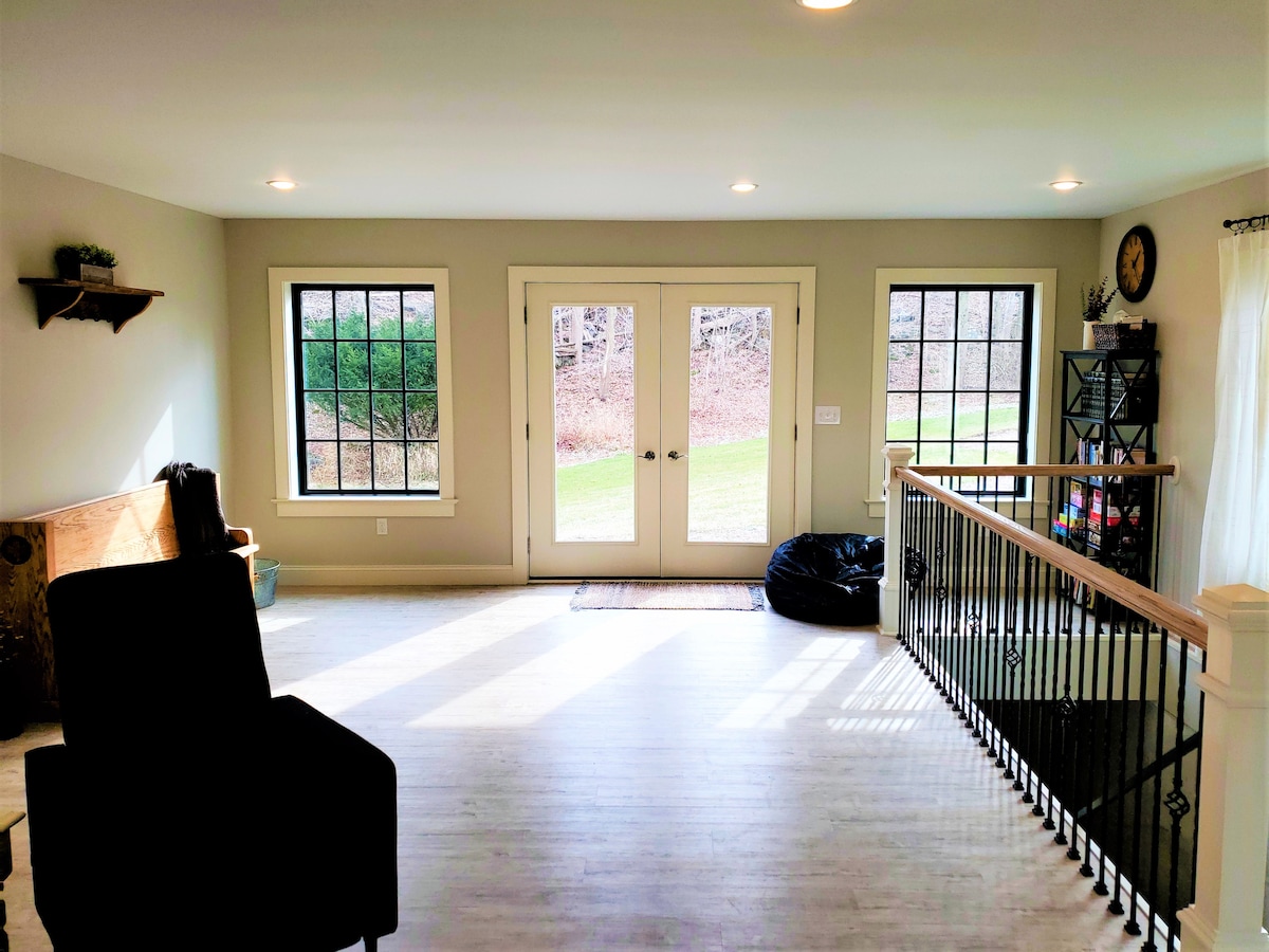 A spacious loft area is illuminated by natural light entering through multiple windows. The room features a comfortable chair, a black bean bag, and a decorative wall shelf. Double doors open to greenery outside, enhancing the open atmosphere.