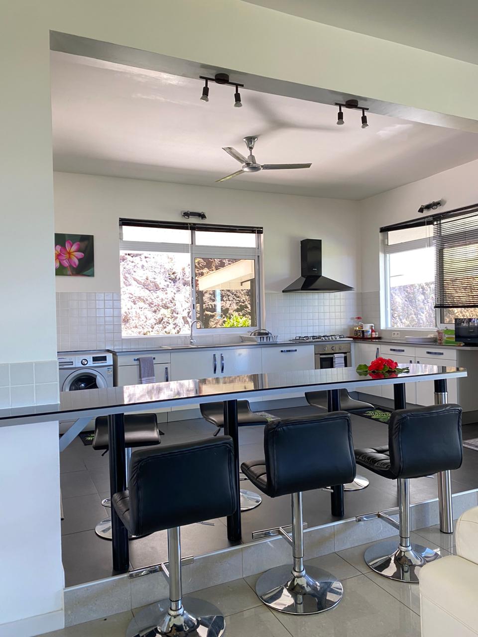 The kitchen features modern appliances, including a stove and washing machine, situated against clean white cabinetry. A sleek bar area with four black stools allows for casual dining, while large windows provide natural light and a view of the surroundings.