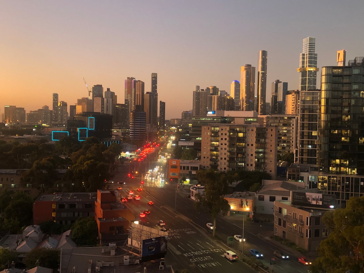 A city skyline is visible at dusk, illuminated by city lights and the glow of sunset colors. Traffic moves along a busy road in the foreground, while modern high-rise buildings and residential structures dominate the background.