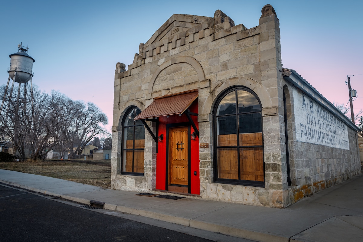 The historic building features a unique stone faƧade, highlighted by arched windows with wooden shutters. A vibrant red door adds a pop of color, inviting guests into the Scandinavian Hall. A quiet street and water tower can be seen in the background.