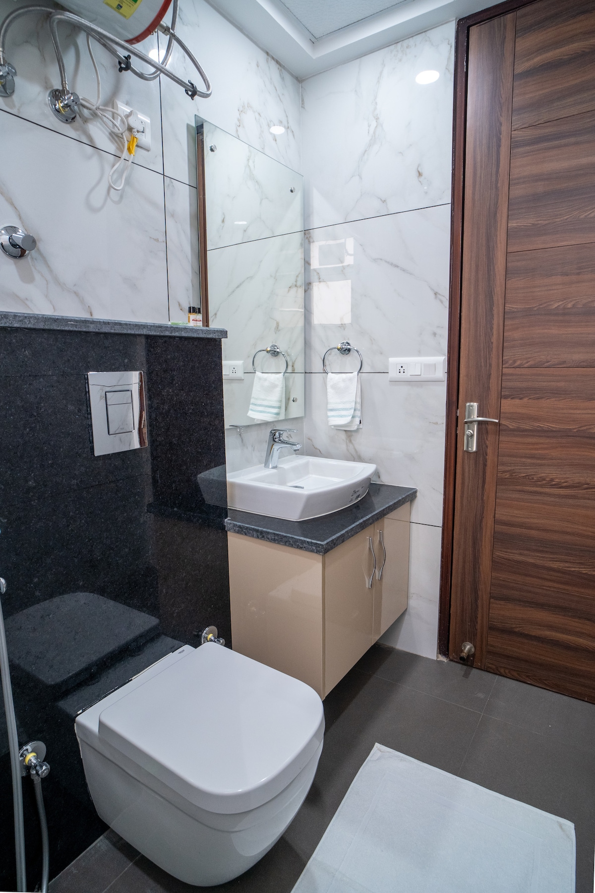 A modern bathroom is depicted featuring sleek, white marble walls and a dark gray countertop. A white toilet is positioned beside a washbasin with a mirror above, while fresh towels are neatly arranged on wall-mounted fixtures. The door showcases a rich wooden finish.