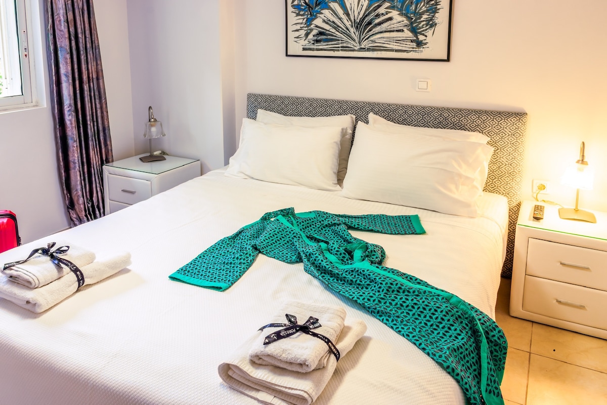 A spacious sleeping area is presented with a large bed featuring crisp white linens. A light blue robe is draped casually over the bed. Two neatly folded towels are placed at the foot of the bed. Soft lighting is provided by bedside lamps and natural light enters through a window.