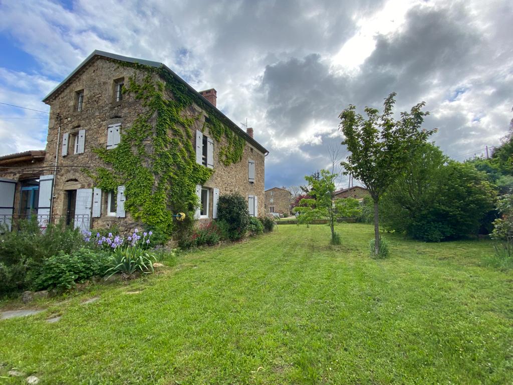 The rustic stone house features climbing greenery along its exterior. The surrounding grassy area is dotted with blooming flowers and trees, while a cloudy sky adds depth to the scene. The inviting yard offers ample space for relaxation and outdoor activities.