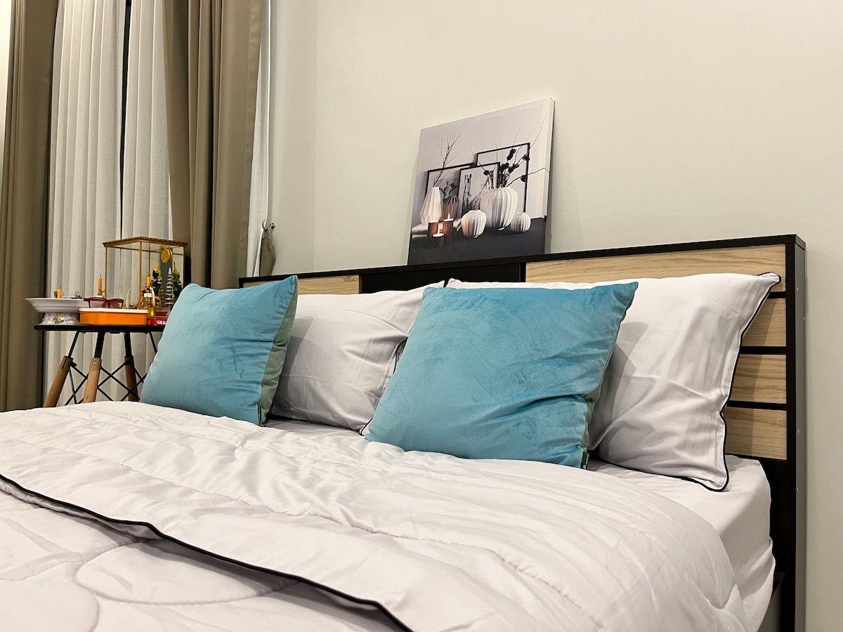 A spacious bed is shown with light-colored bedding featuring a subtle pattern. Two plush blue pillows are arranged at the headboard. A decorative wall art piece is displayed above the bed, with a small side table adjacent, holding ornamental items.