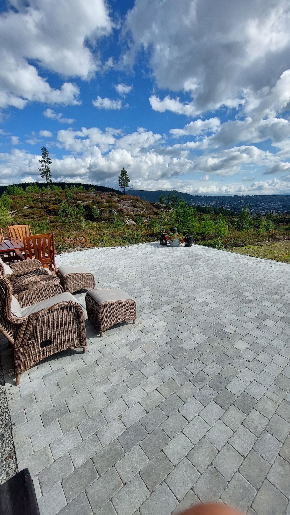 A spacious outdoor patio area features patterned stone flooring, surrounded by natural landscape. Comfortable seating options are provided, including wicker chairs and benches. The scenic view showcases rolling hills under a partly cloudy sky, emphasizing a serene and inviting environment for relaxation.