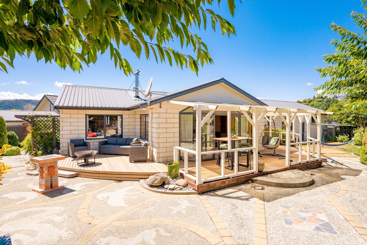 The exterior of a single-story home is presented, featuring a large deck with outdoor seating. Surrounding greenery adds to the peaceful ambiance. A stone pathway leads to a decorative stone area with a fire pit, highlighting the blend of indoor-outdoor living.