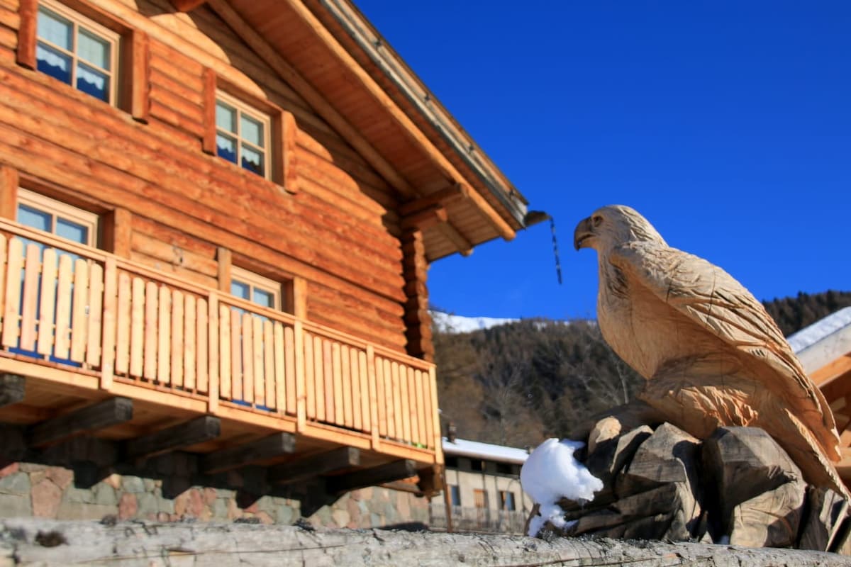 A traditional wooden chalet features a charming carved eagle statue positioned on a stone base in front. The building showcases a balcony with multiple wooden railings and large windows, complemented by snow-capped mountains in the background.