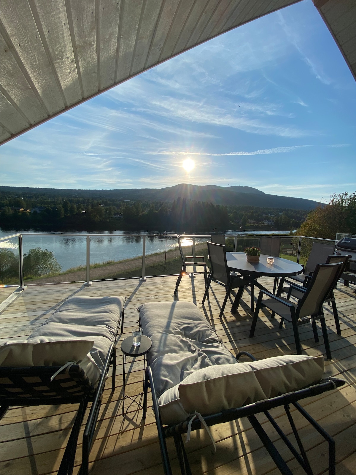 A spacious terrace offers a relaxing seating area with lounge chairs and a dining table, overlooking a scenic river and distant hills. Sunlight bathes the space, creating a serene atmosphere while the surrounding landscape is enhanced by natural beauty.
