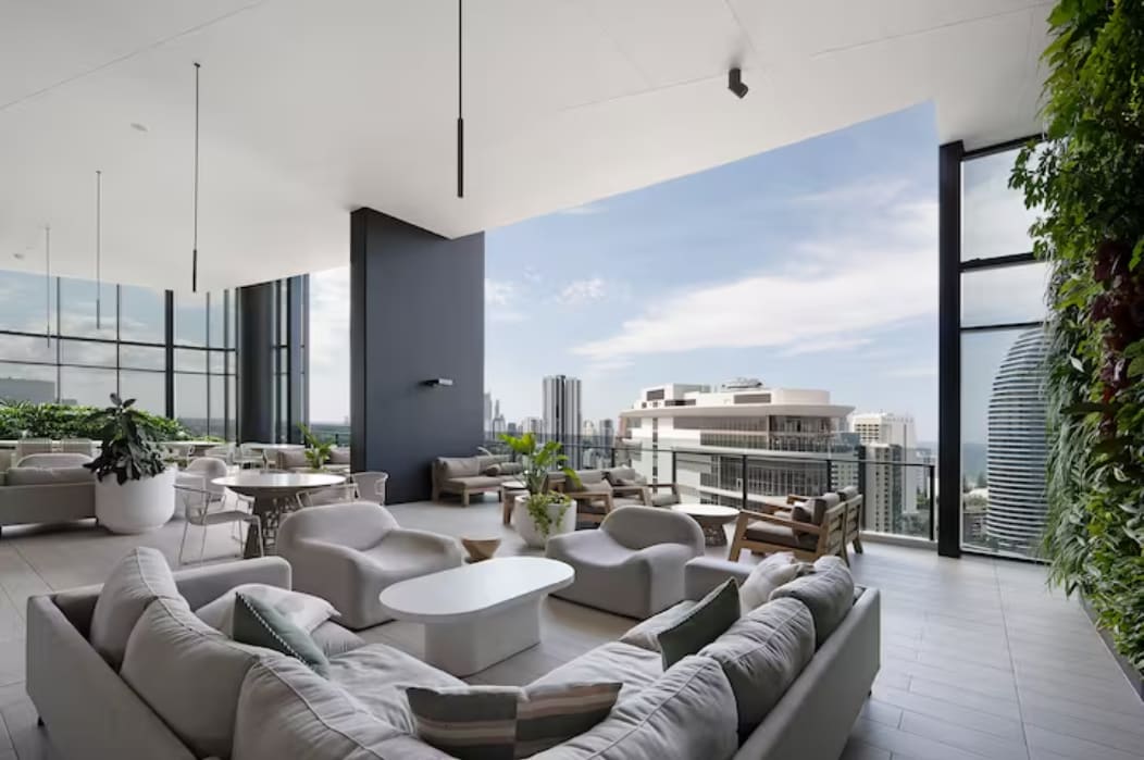 An expansive common area features modern furniture arranged for relaxation and socializing. Large glass windows offer unobstructed views of the skyline and ocean beyond. Lush greenery is incorporated into the design, enhancing the inviting atmosphere while contributing to a sense of tranquility.