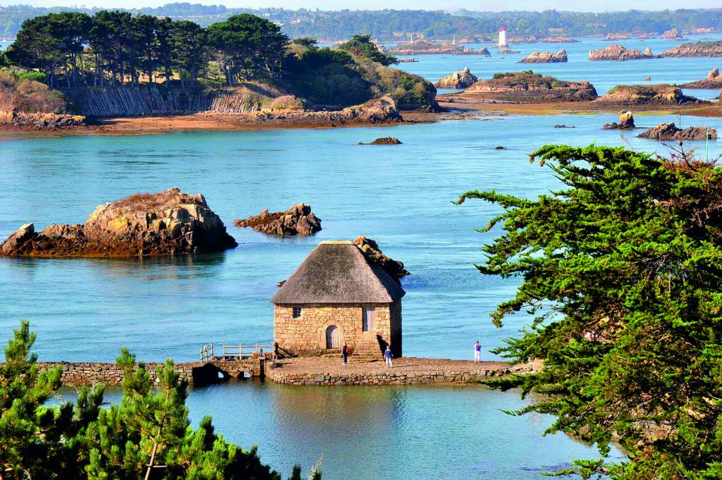 A quaint stone building with a thatched roof is situated on a rocky shore, surrounded by calm waters. Nearby, natural rocky formations emerge from the sea, with greenery lining the coastline. A lighthouse stands in the distance, completing the serene coastal landscape.