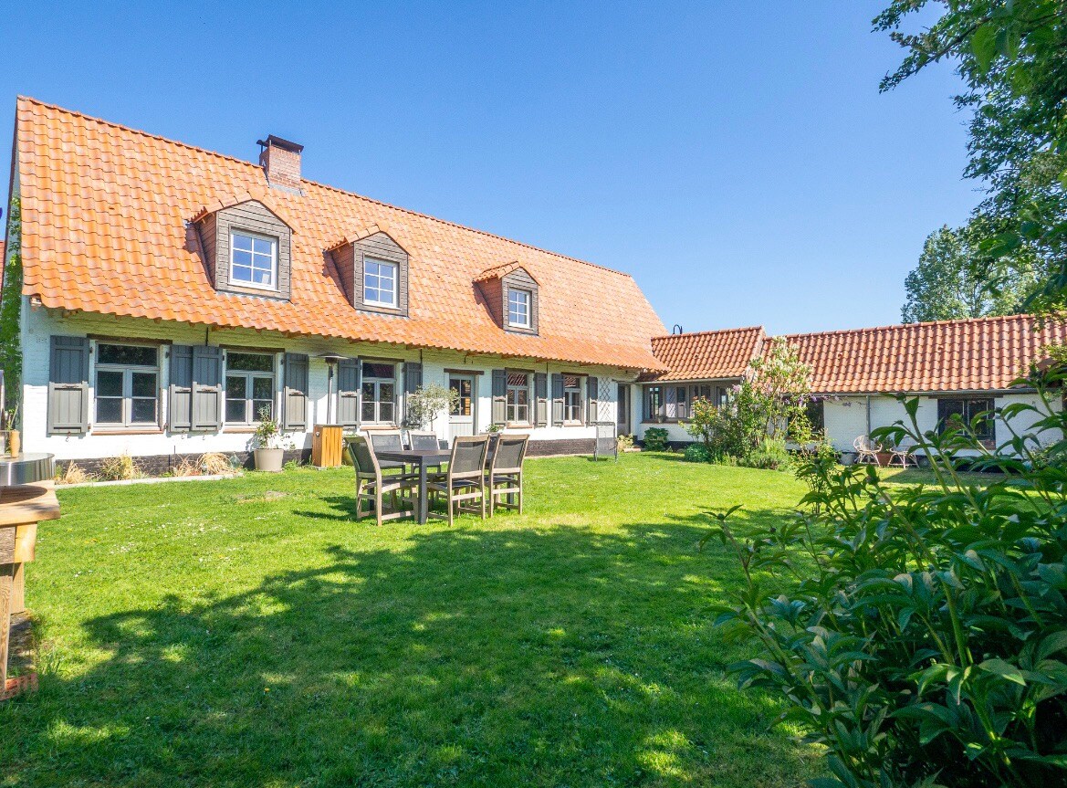 A large home with a traditional roof is surrounded by a spacious green lawn. Outdoor dining furniture is arranged on the grass, offering a place for guests to gather. The façade features multiple windows with shutters, enhancing the building's character.