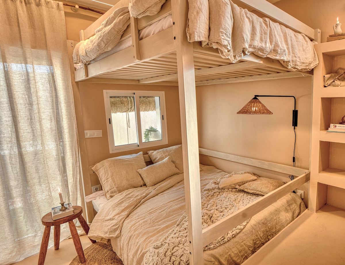 An open sleeping area features a spacious double bed beneath a lofted bed frame. Soft neutral tones are present in the bedding and decor, complemented by natural light filtering through a nearby window. A small wooden table with a candle adds a cozy touch.