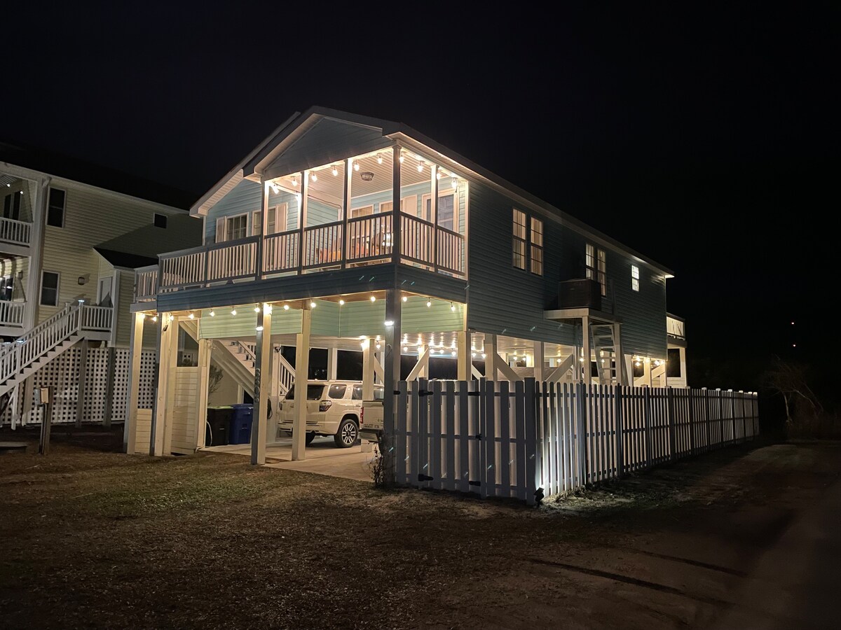 The exterior of the two-story home is illuminated by soft lighting at night, highlighting the spacious front deck and multiple entry points. A fenced yard surrounds the property, with a parking area visible beneath the house. The structure blends seamlessly with the surrounding landscape.