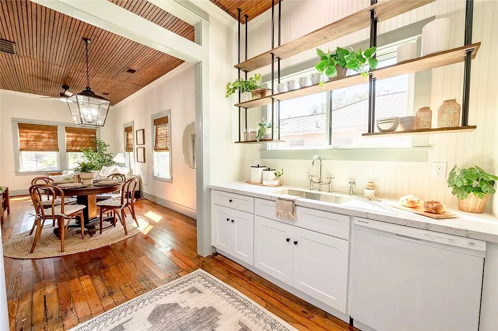 A bright and inviting kitchen area is displayed, featuring open shelving adorned with greenery. The cabinetry is complemented by a marble countertop, while a dining space with a round table and rattan chairs is visible in the background, illuminated by natural light.