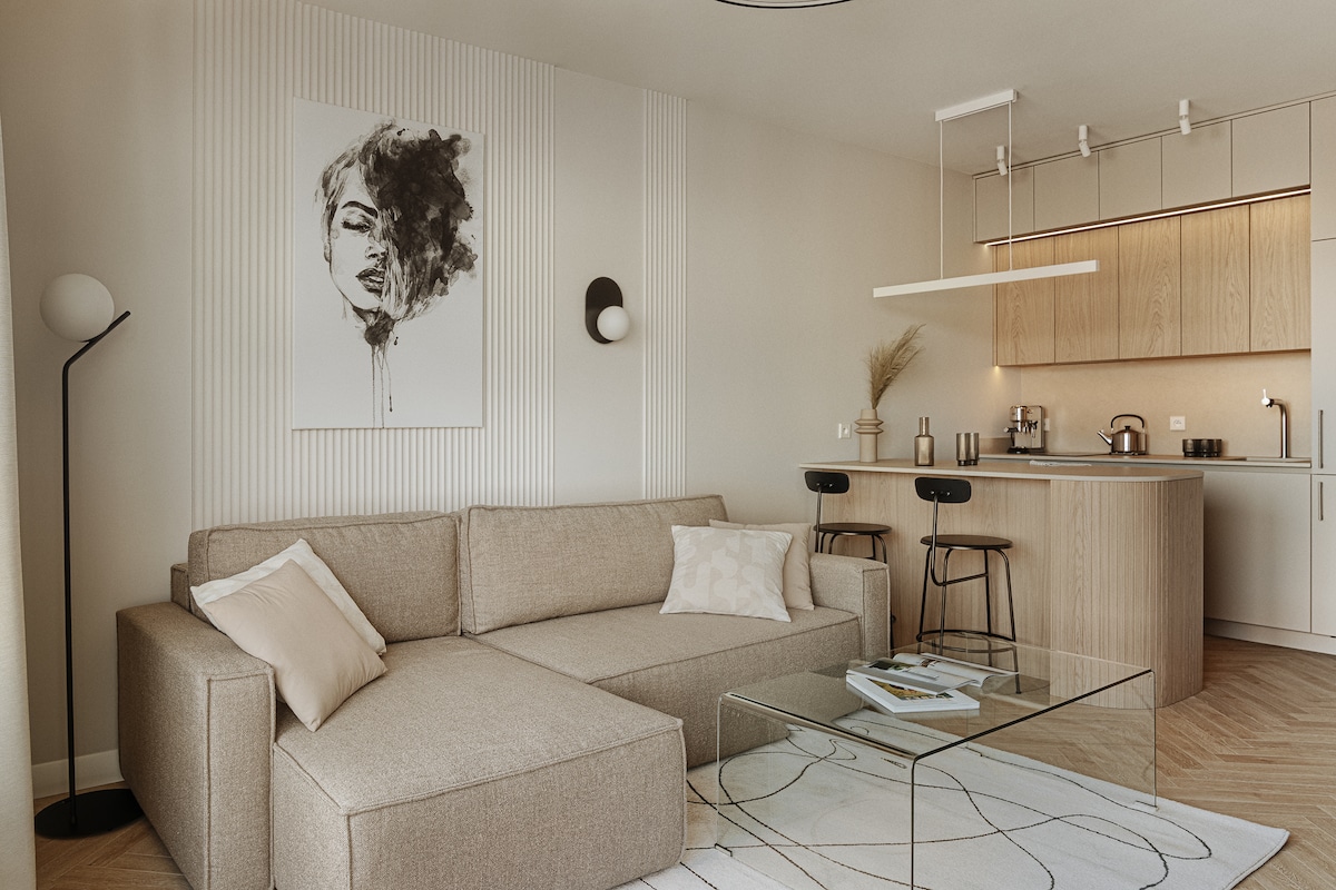 A modern living area features a spacious beige sofa with an accompanying decorative pillow. A light wood kitchen area is visible in the background, complemented by a glass coffee table and minimalist decor. Soft lighting enhances the calming atmosphere.