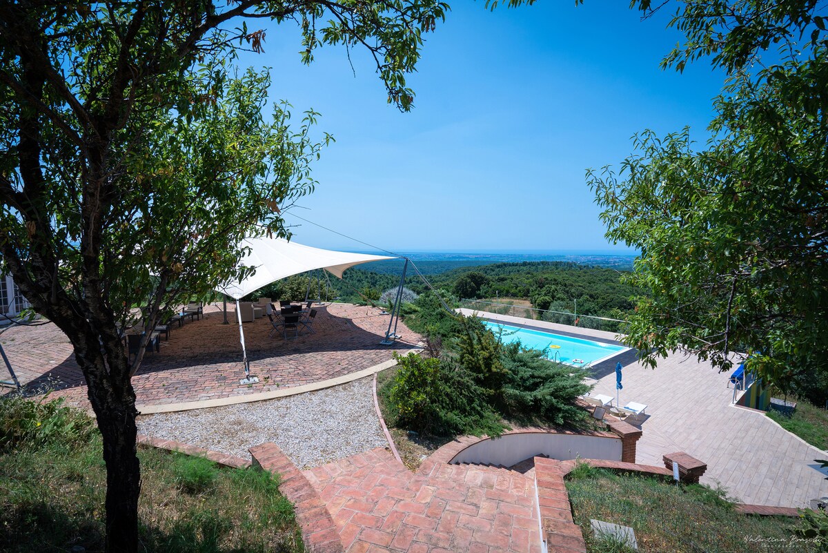 The outdoor area features a shaded seating space under a large canopy, offering views of a swimming pool and expansive greenery beyond. A pathway made of brick steps leads down to the relaxing pool area, framed by trees and lush vegetation.