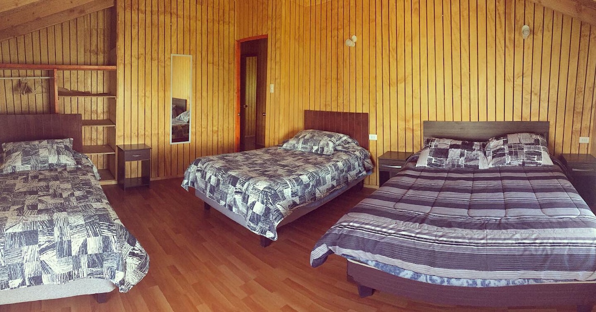 A spacious bedroom features three large beds, each adorned with patterned bedding. Warm wooden walls enhance the cozy atmosphere. A shelf and a mirror contribute to the room's functionality, while natural light enters through a nearby door.