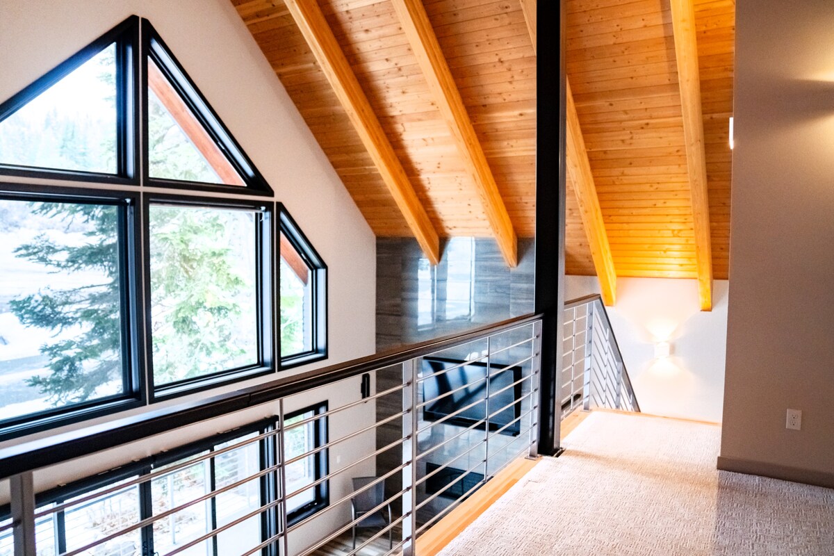 The loft area features vaulted wooden ceilings with exposed beams and large windows that invite natural light. A balcony overlooks the lower levels, providing views of the surrounding trees. The space is designed with modern finishes and a neutral color palette.