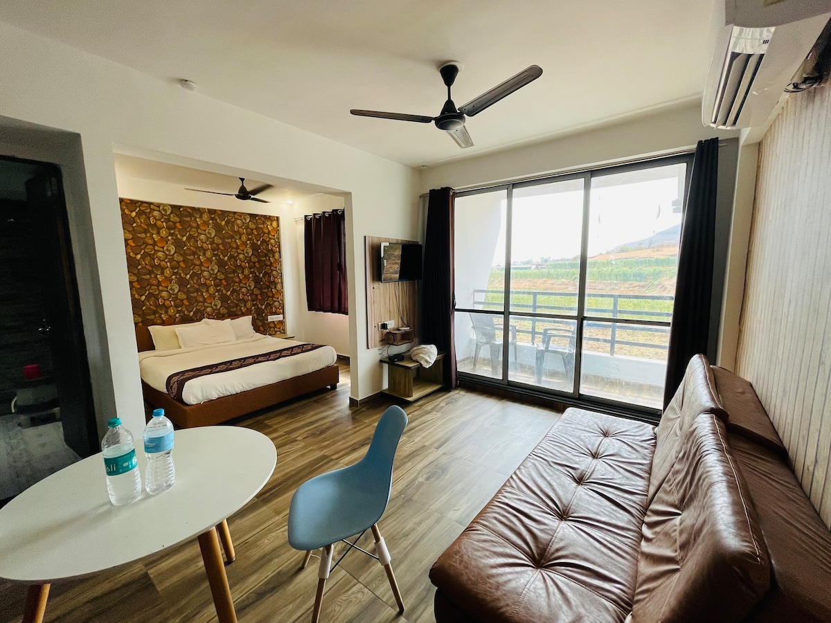 A spacious hotel room features a large bed with a supportive headboard and a patterned accent wall. A comfortable seating area includes a brown sofa and a round dining table with two water bottles. Floor-to-ceiling windows provide natural light and views of the surrounding landscape.