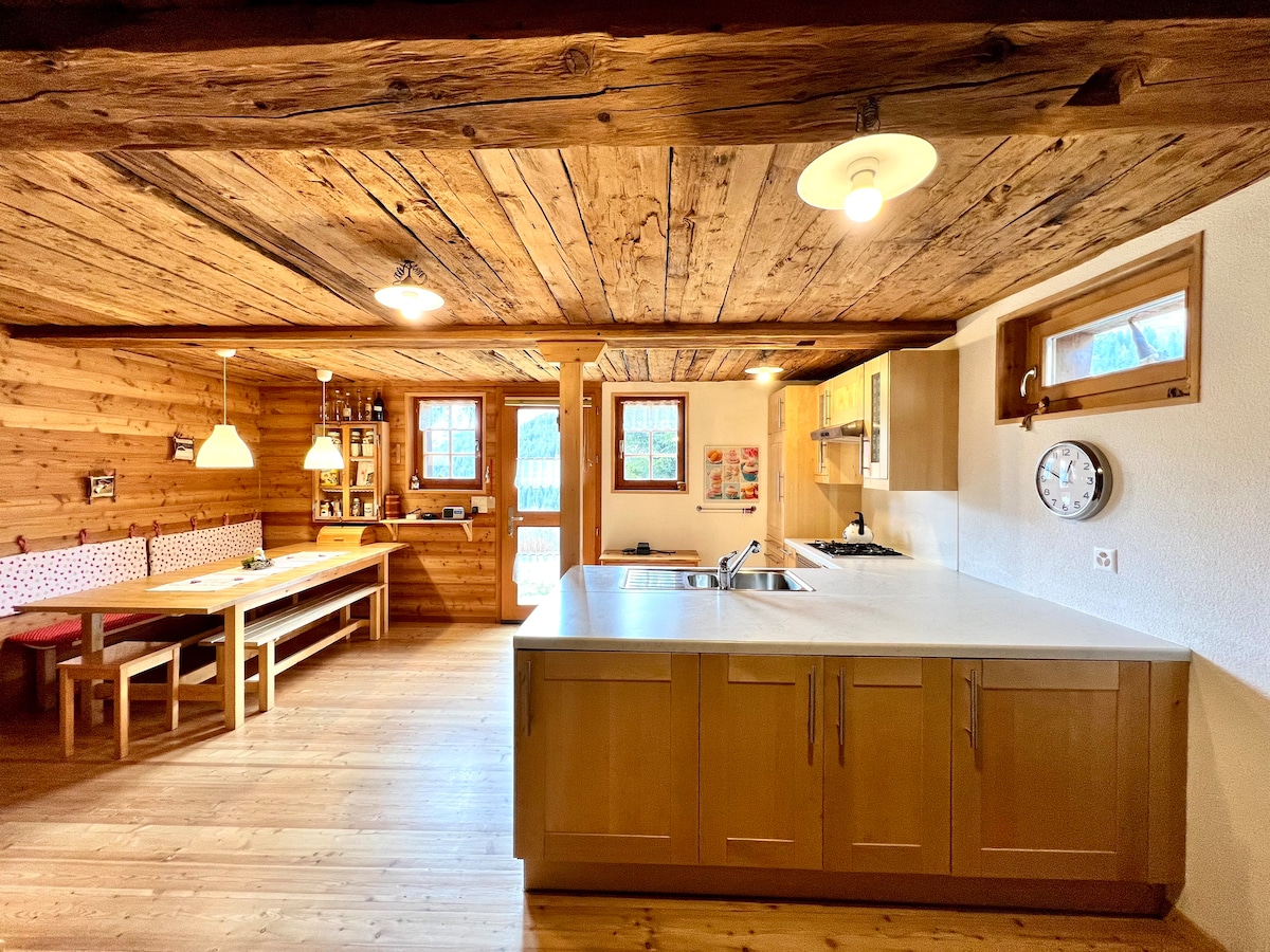 A spacious and well-equipped kitchen is visible, featuring wooden cabinetry and a large island. Natural light enters through windows, illuminating the rustic wooden beams and walls. A dining area with a long table and benches is situated nearby, enhancing a communal atmosphere.