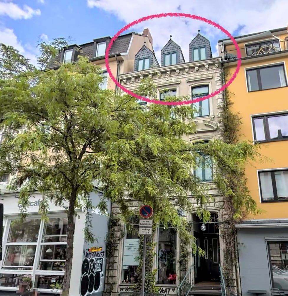 The historic building features a classic façade adorned with architectural details. The roof showcases distinctive pointed dormers, adding character to the structure. Surrounding trees provide greenery, while the street below reflects a vibrant urban atmosphere with various shops and cafés.