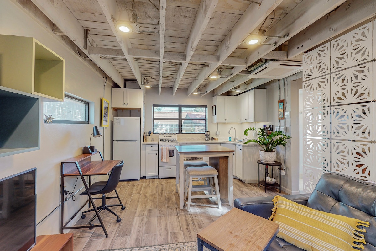An open studio space is showcased with wooden beams above and natural light filtering through a window. The kitchen features white cabinetry, a gas stove, and a dining area with a two-person table. A comfortable sofa and a small desk complete the inviting layout.