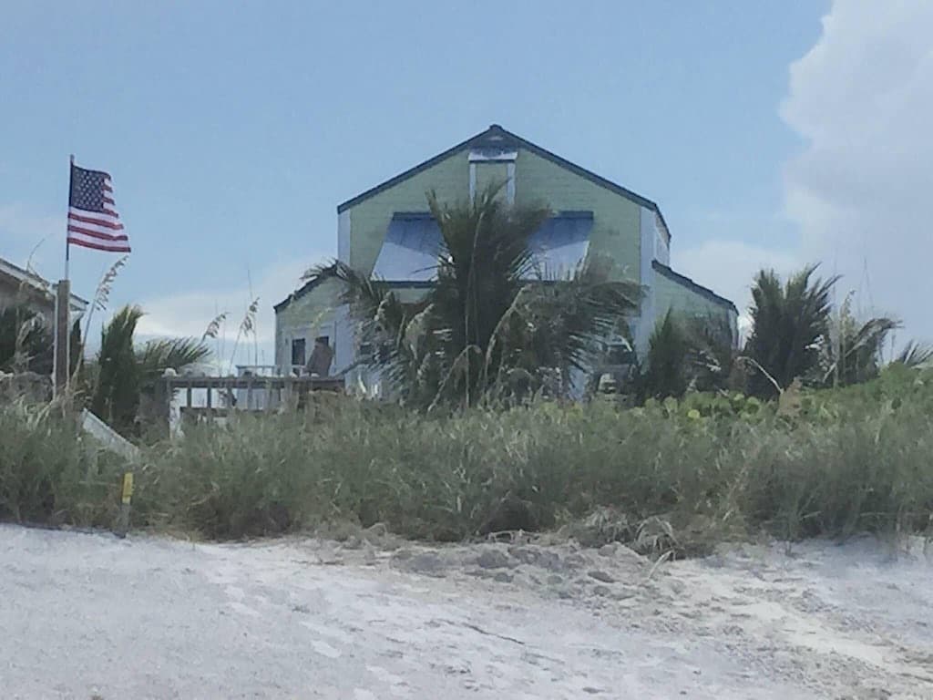 A beach home is set against a backdrop of sandy terrain and green foliage, featuring a prominent American flag on display. The structure is painted in a light green hue, with blue accents complementing its coastal appeal.