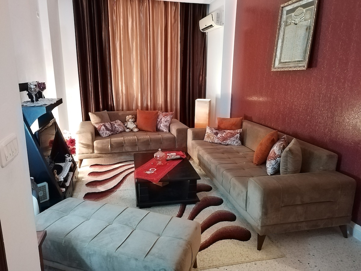 A comfortable living area features a large, L-shaped beige sofa adorned with patterned cushions. A dark coffee table sits in the center, surrounded by a plush, multi-patterned rug. Drapes in rich shades frame a window, enhancing the warm ambiance of the room.