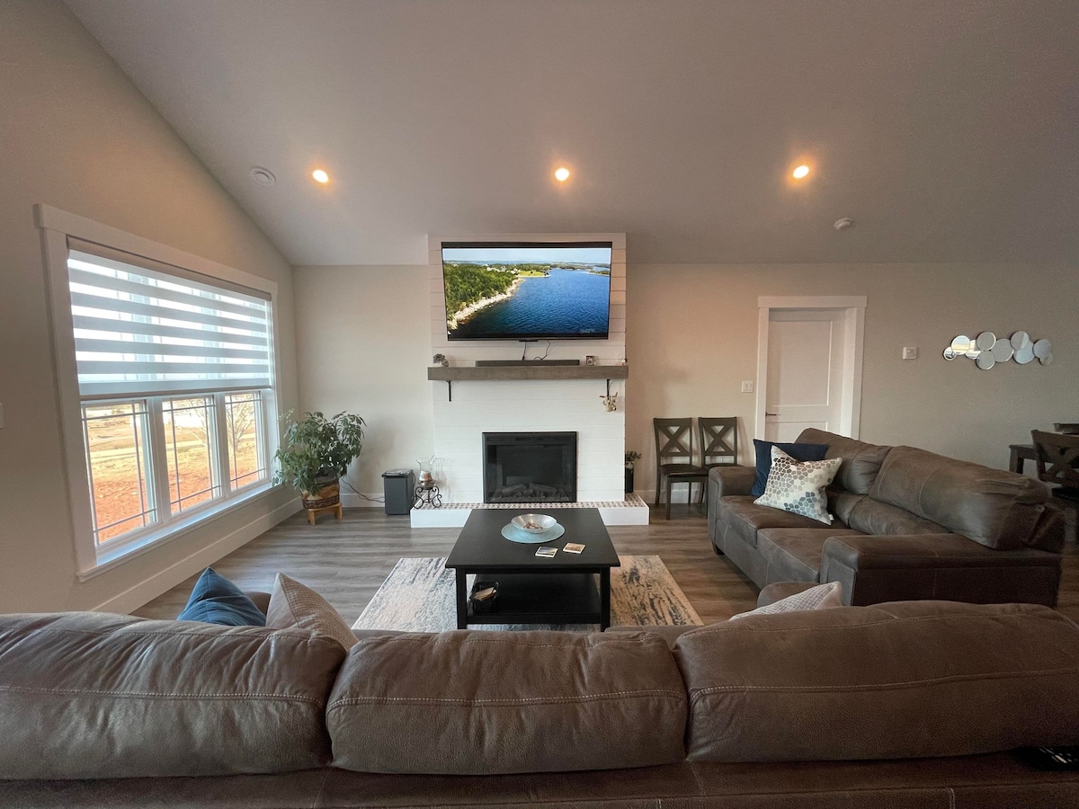 The living area is furnished with a spacious sectional sofa and two armchairs, complemented by a coffee table at the center. A large television is mounted above the fireplace. Natural light filters through the window adorned with blinds, illuminating the room.