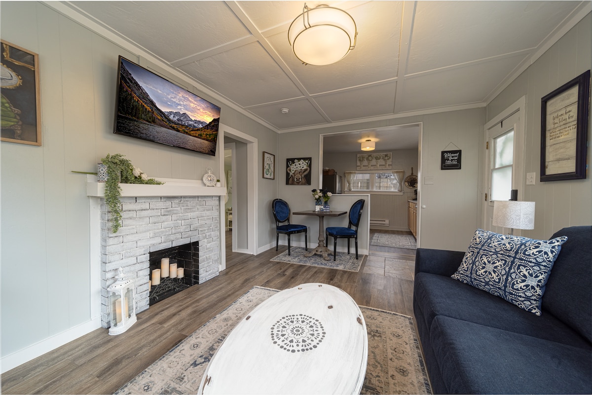 The living area features a dark blue sofa facing a whitewashed brick fireplace, where candles are arranged. A round coffee table with a decorative design sits in front. A mounted TV displays a scenic mountain view, while a dining area with two chairs is seen in the background.