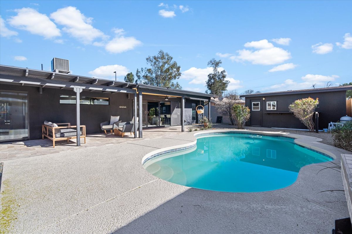 An outdoor space features a uniquely shaped swimming pool surrounded by a light-colored concrete patio. Lounge seating is positioned nearby under a covered area. Lush greenery is evident along the perimeter, accompanied by a clear blue sky overhead.