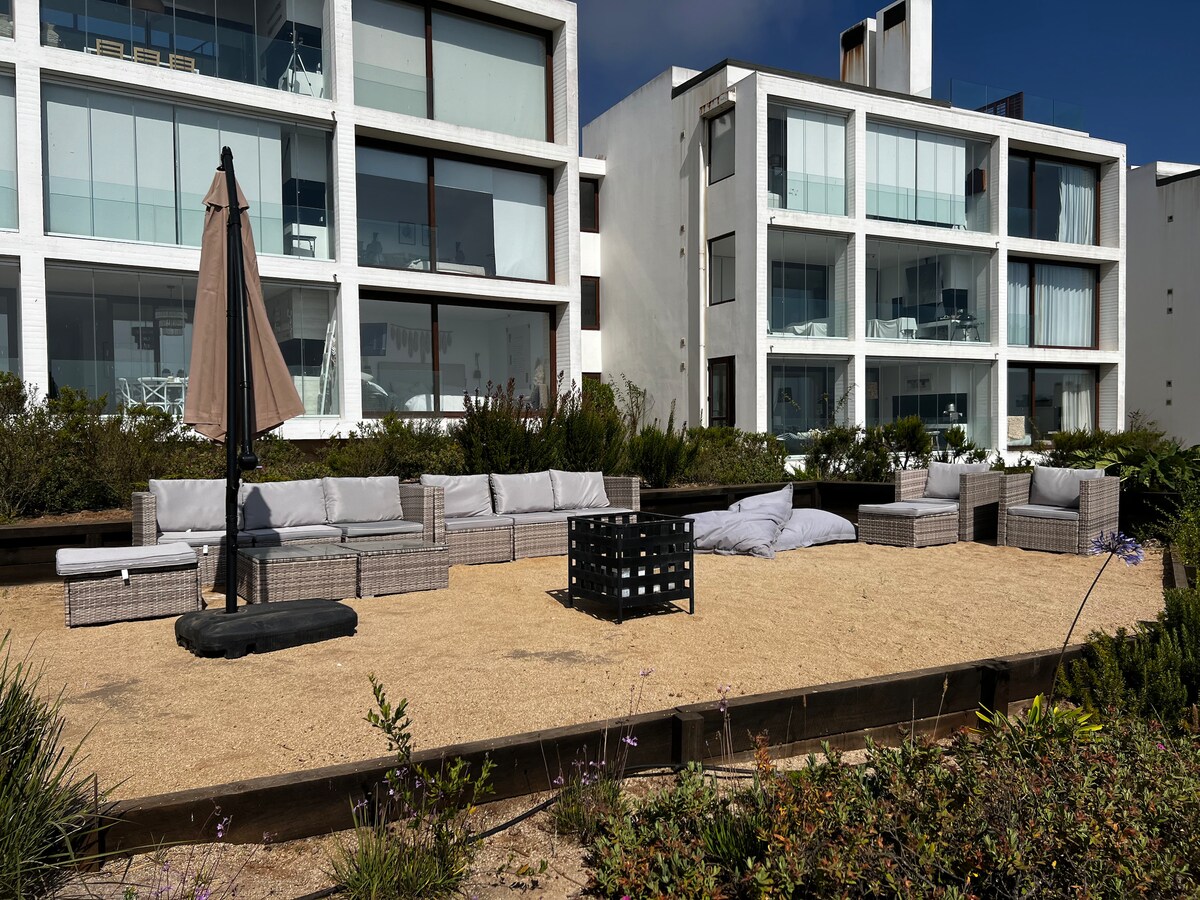 An outdoor area features modern lounge seating arrangements with light grey cushions and a central black fire pit. Surrounding vegetation is present, and a large umbrella is positioned to provide shade. The backdrop includes contemporary buildings with large windows reflecting natural light.