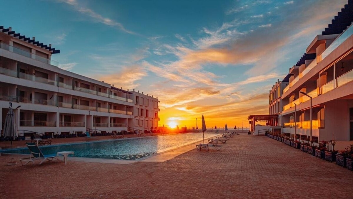 A stunning sunset is captured over a tranquil outdoor swimming pool, framed by two modern apartment buildings. Sun loungers are positioned alongside the pool area, and the warm tones of the sky are reflected on the water's surface.