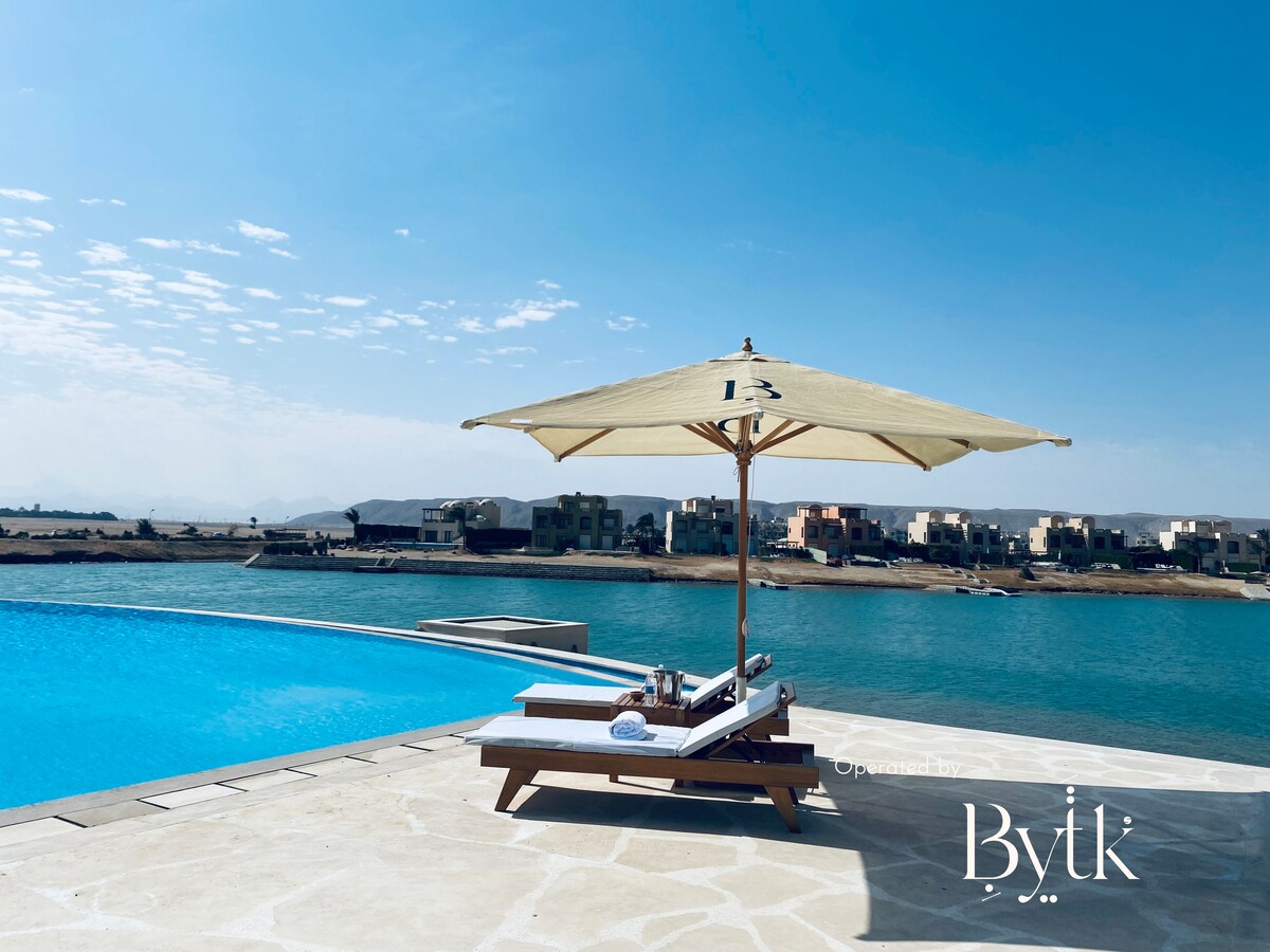 A sun lounger with a beach umbrella is set beside a refreshing swimming pool, overlooking a serene lagoon. The landscape beyond features a row of buildings against clear blue skies, creating a peaceful atmosphere.