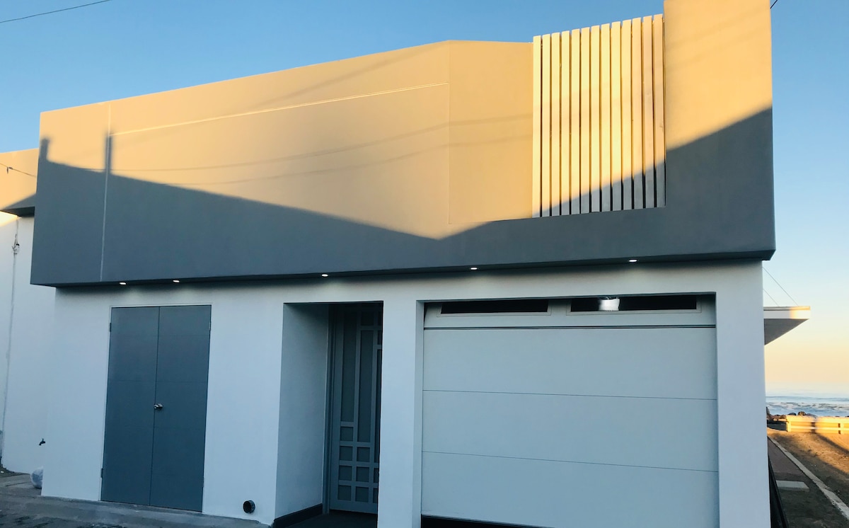The exterior of the house is shown, featuring a modern design with a neutral color palette. The front door is framed by a decorative panel, and a garage door is visible. Soft shadows from the building's architecture emphasize the clean lines and simple structure.