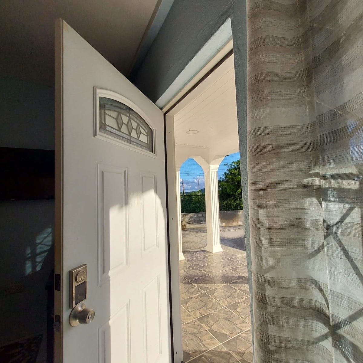 The open door invites guests into the home, revealing a spacious patio area with arched columns. Natural light brightens the entry, where a patterned floor enhances a sense of openness. The gentle folds of the curtain add a touch of softness to the entrance.