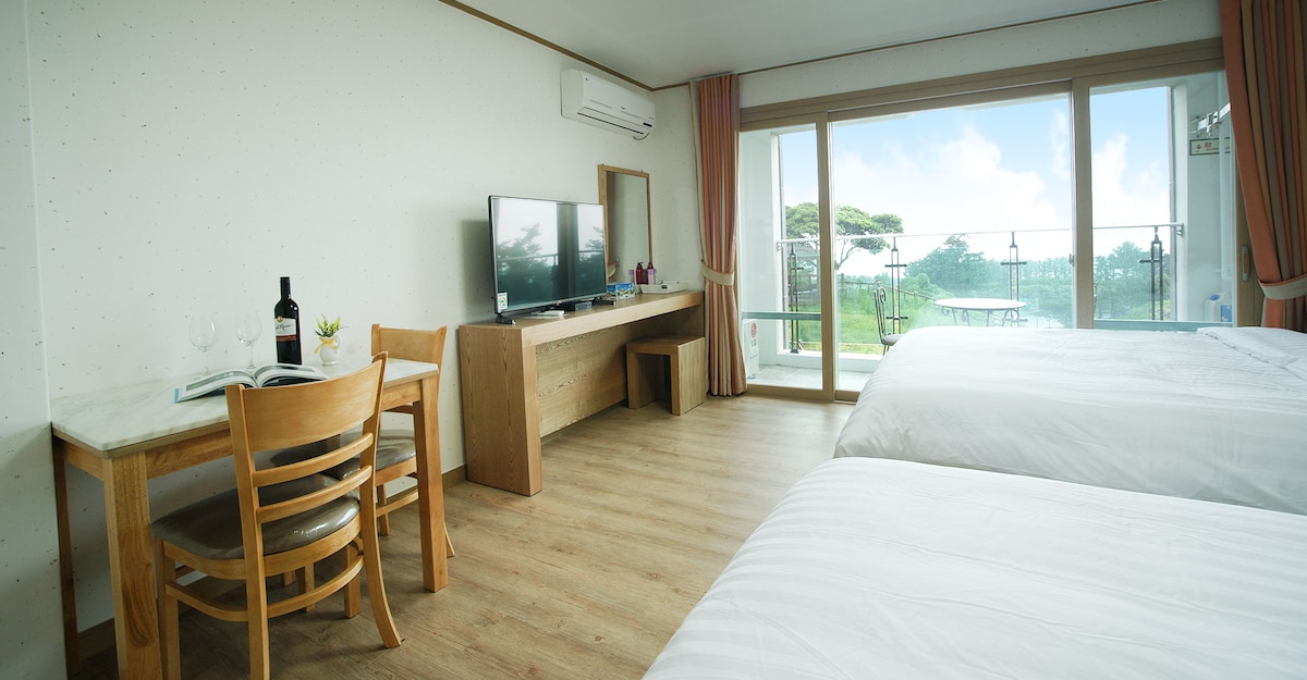 A bright room features two spacious beds and a large glass door leading to a balcony. A television is placed on a wooden console, while a small dining table with chairs is positioned nearby. Natural light floods the space, creating a welcoming atmosphere.