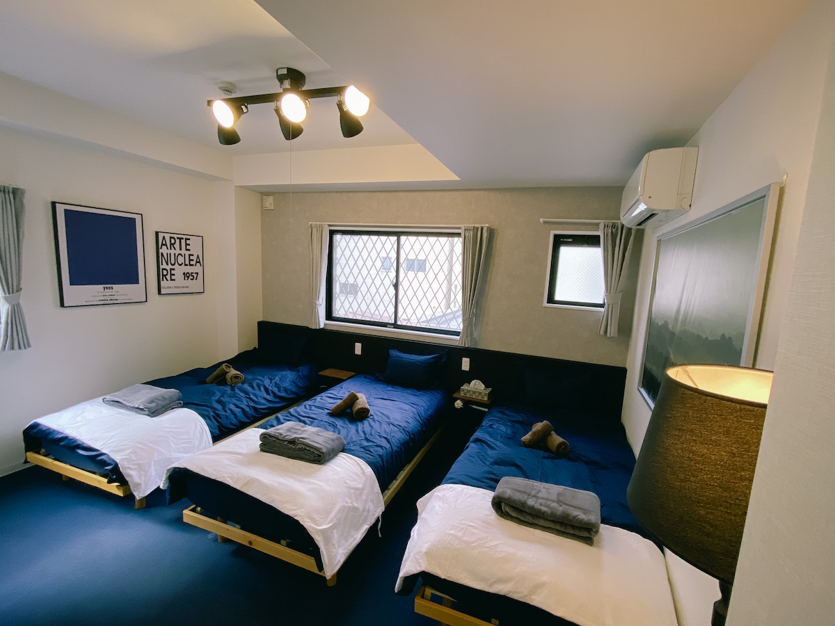 The room features three single beds positioned on wooden platforms, adorned with navy blue bed linens. A large window with a grid pattern allows natural light to enter, complemented by curtains. A lamp stands beside one bed, providing additional lighting, and a minimalistic design is evident throughout.