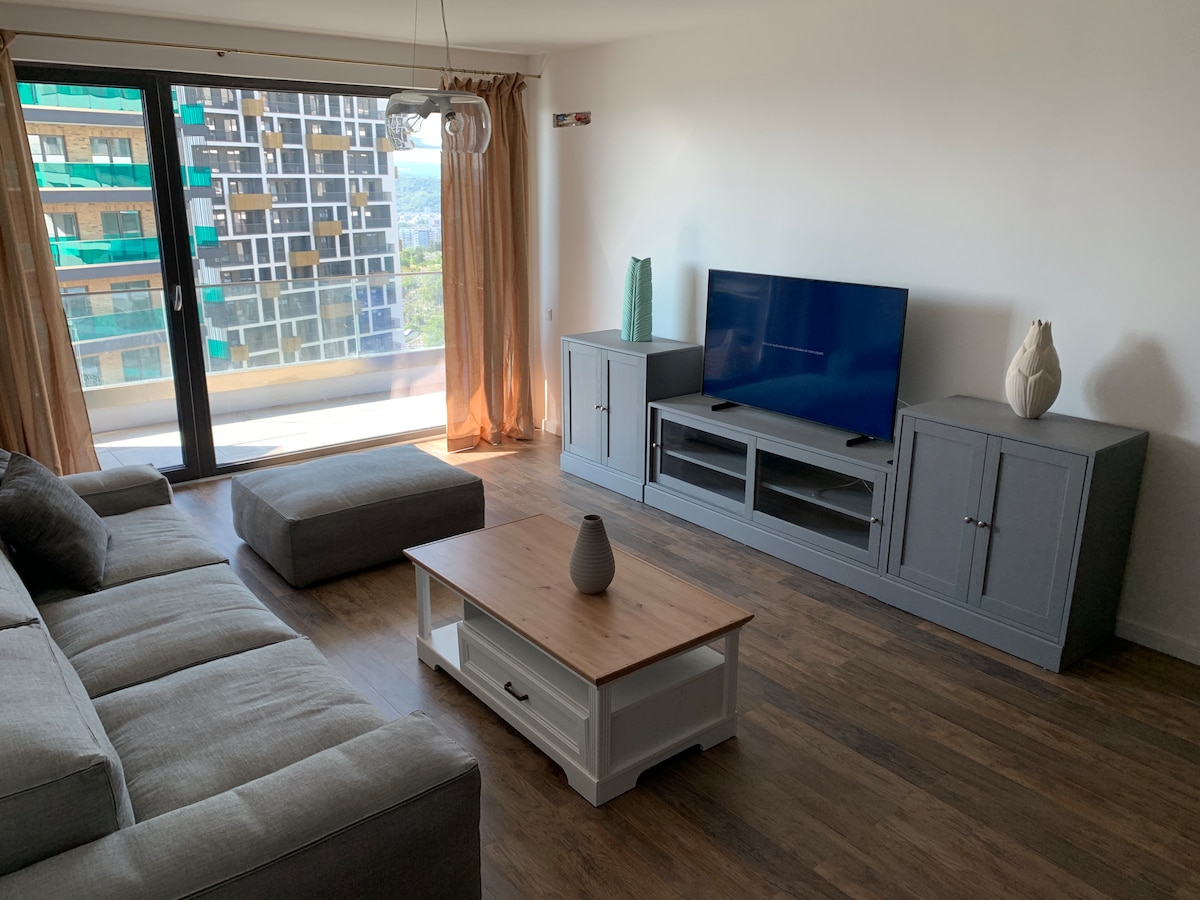 A modern living area features a spacious grey sectional sofa and a plush ottoman. A wooden coffee table sits in front, complemented by a television mounted on a grey media console. Large glass doors lead to a balcony, allowing natural light to fill the space.