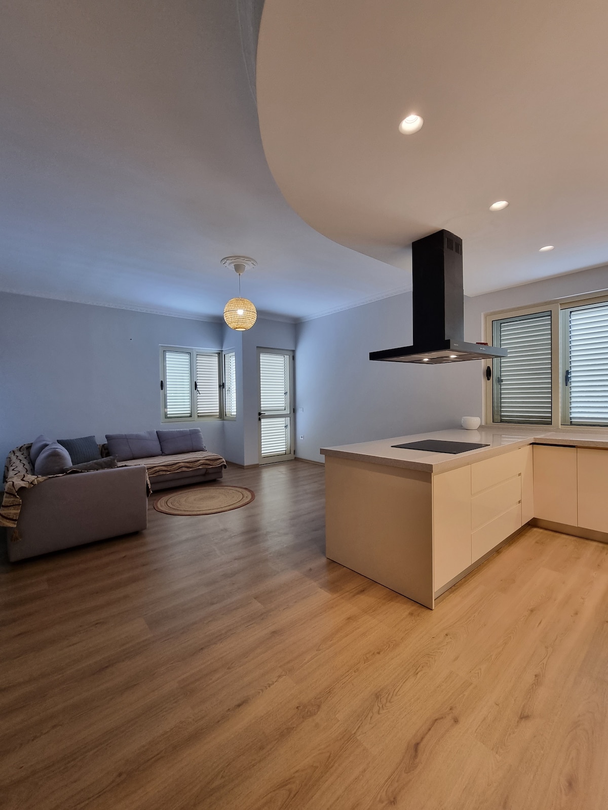 An open living space features a modern kitchen with an island, complemented by a sleek range hood. A comfortable sofa is positioned against a light blue wall, with natural light streaming in through multiple windows. Warm wood flooring adds to the inviting environment.