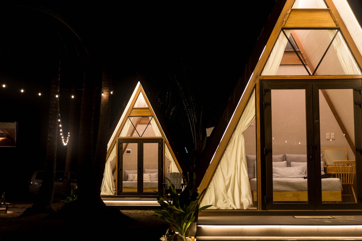 Two A-frame structures are illuminated by soft lighting at night, highlighting their wooden features and large glass doors. Natural shades and warm light create a welcoming atmosphere, while palm trees frame the scene, emphasizing the tranquil setting.