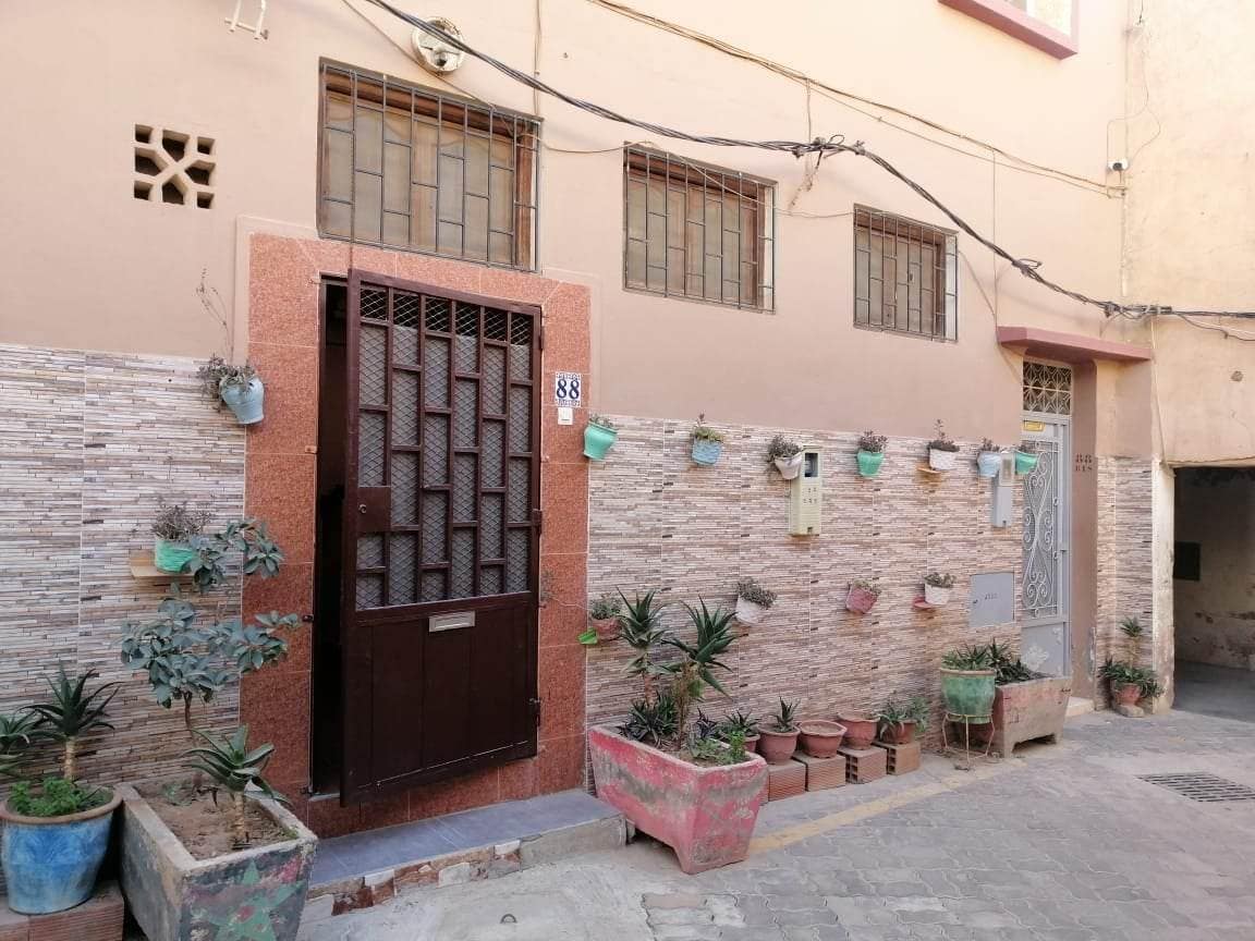 The exterior of a duplex is presented, showcasing a textured stone wall with a neatly framed wooden door. Potted plants line the entry, adding greenery. Windows are visible above the entrance, enhancing natural light exposure.