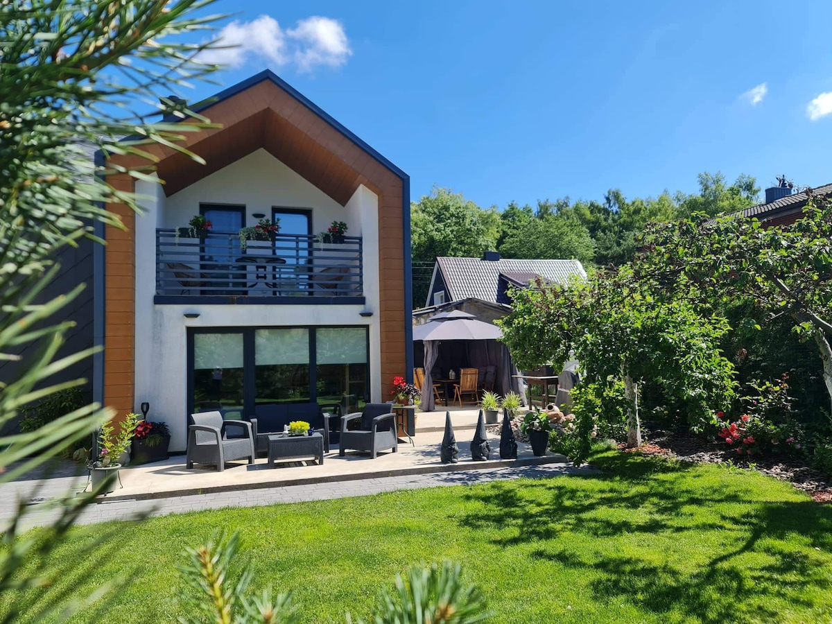 The exterior of a well-maintained house is shown, featuring a small patio area with seating and an umbrella. Lush greenery surrounds the property, with colorful plants and trees contributing to the tranquil environment. A clear blue sky adds to the inviting atmosphere.