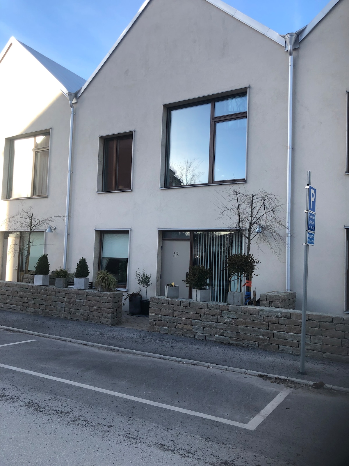 The exterior of a modern building is viewed, with large windows allowing natural light to enter. Planters with small trees and shrubs line the entrance, while a stone wall separates the property from the street. Designated parking is visible nearby.