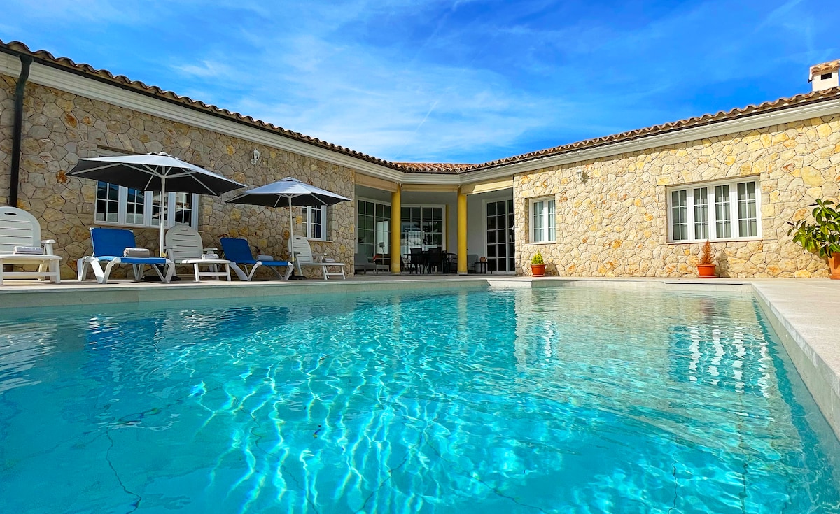 A refreshing swimming pool reflects the blue sky, surrounded by sun loungers and umbrellas. The stone facade of the house provides a rustic charm, with large glass doors opening to the patio area. Natural light illuminates the space, enhancing its serene atmosphere.